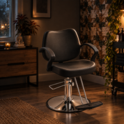 A black salon-style barber chair with chrome base and footrest, shown in a warmly lit room with wood flooring, a window, plants, and wall decor.